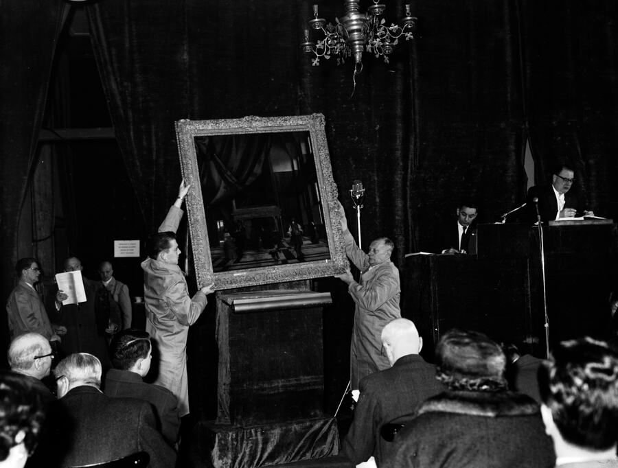 Two porters hold a large gilt-framed painting up to a room of bidders while the auctioneer takes bids, photographed in black and white